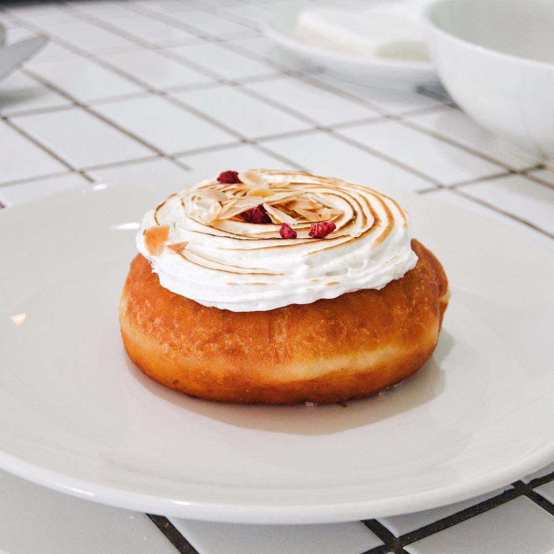 Filled doughnut topped with meringue and raspberry pieces on white plate and tiles.