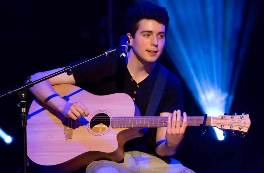 Dark haired young man on stage, blue backlighting, sitting on a chair playing guitar and singing into microphone