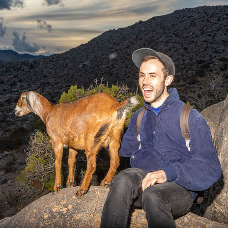 Smiling person sitting next to a brown goat in a desert at sunset.