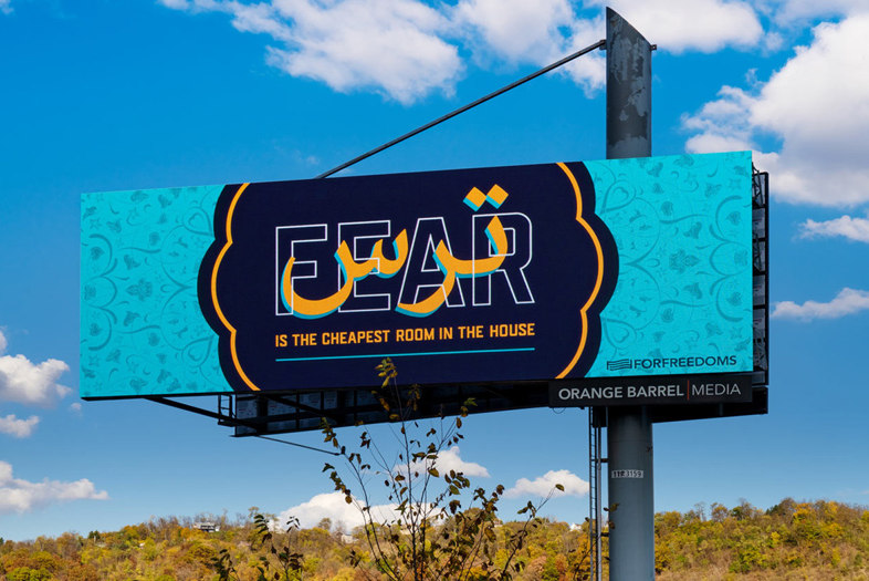 A blue billboard reading &quot;Fear is the cheapest room in the house&quot; photographed in front of a blue sky and autumn trees in the background.