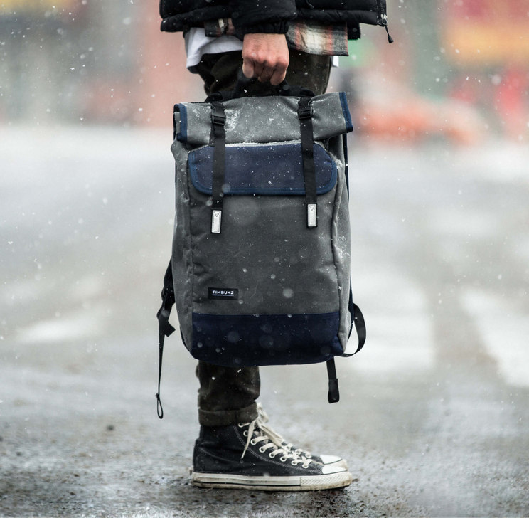 Man holds a Timbuk2 messenger bag featuring custom typographic logo while standing on a street in the snow