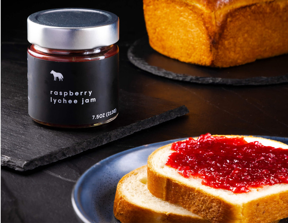 Jam packaging for raspberry lychee jam for craftsman and wolves sits beside a piece of bread spread with jam.